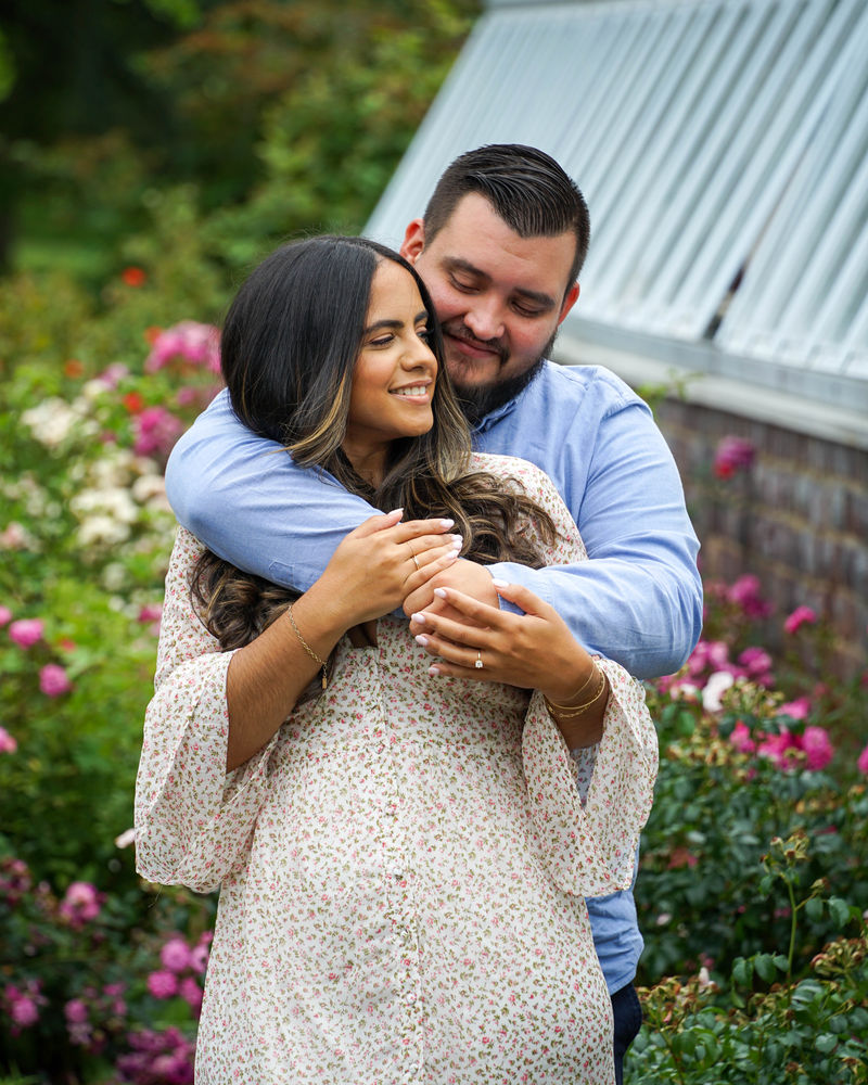 Man embracing his fiancee from behind beside a white garden barn