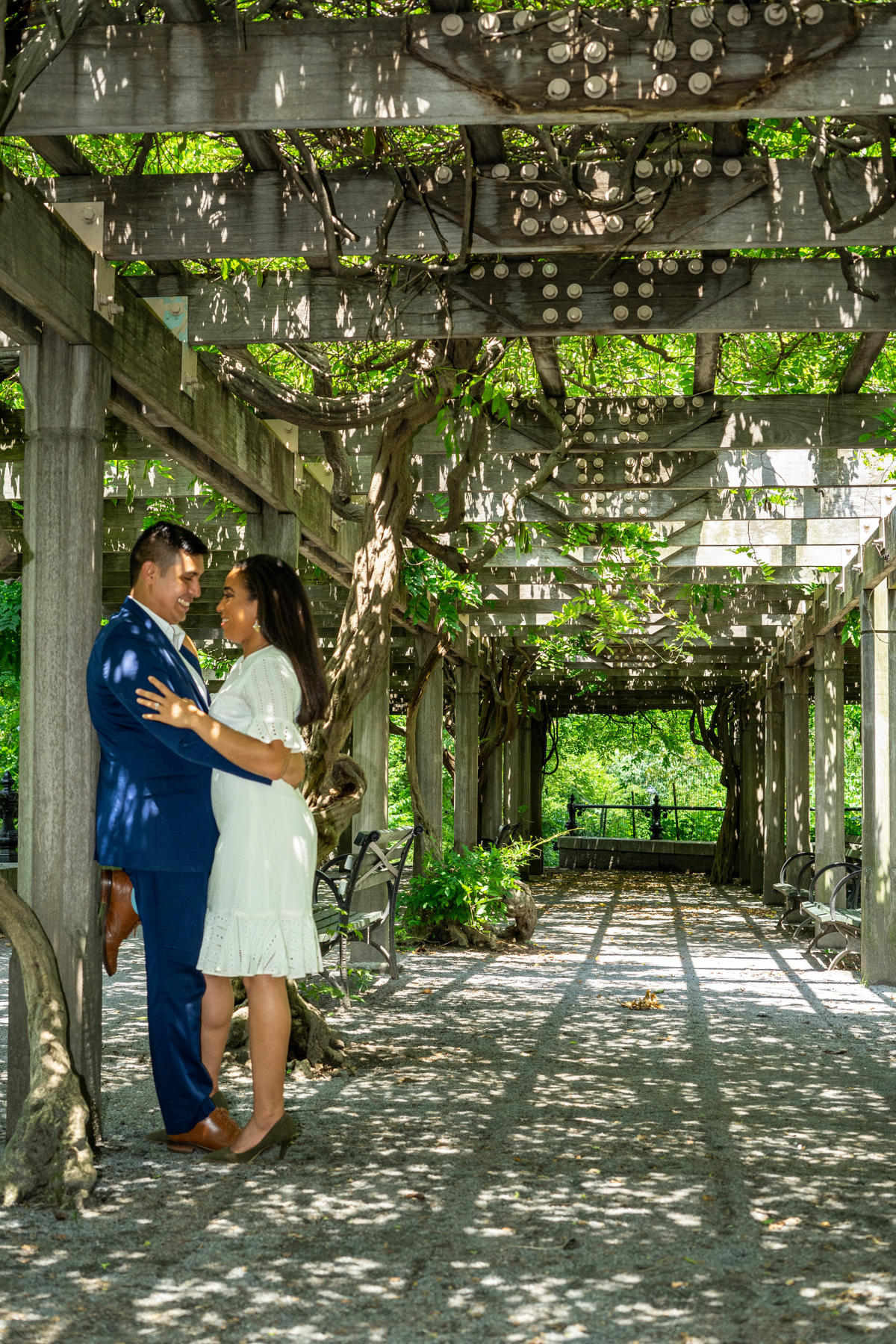 Couple holding each other beneath a sunlit wisteria pergola