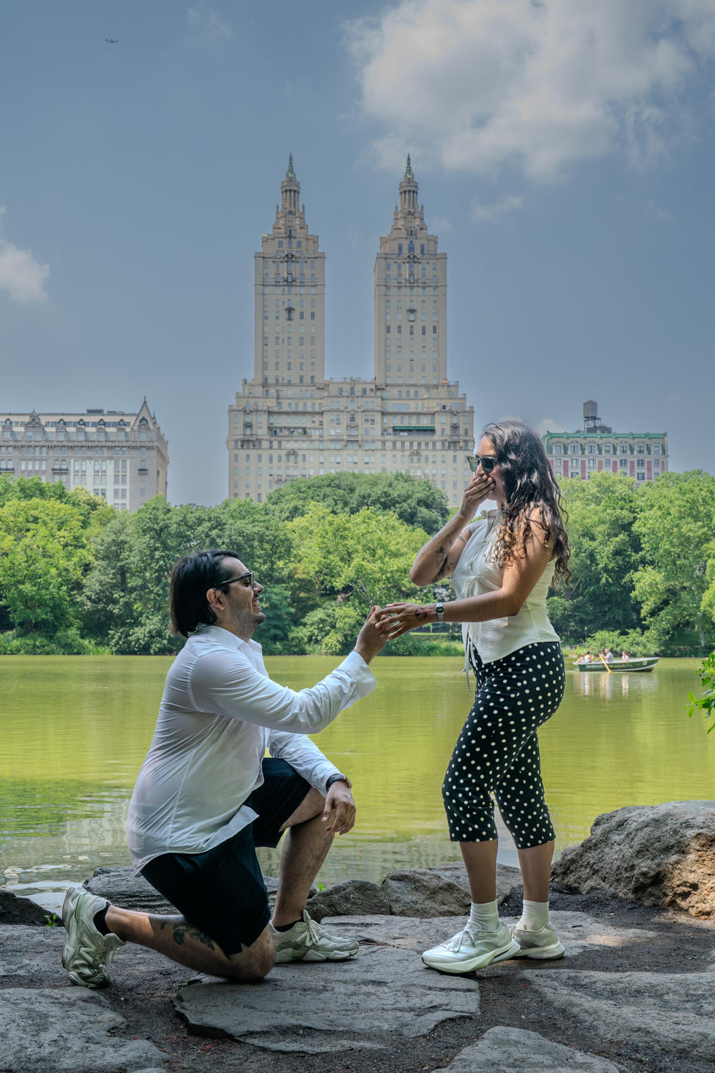Marriage proposal on one knee at the edge of a lake with the city skyline behind