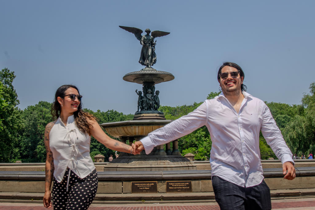 Newly engaged couple reaching for each other's hands in front of a park fountain
