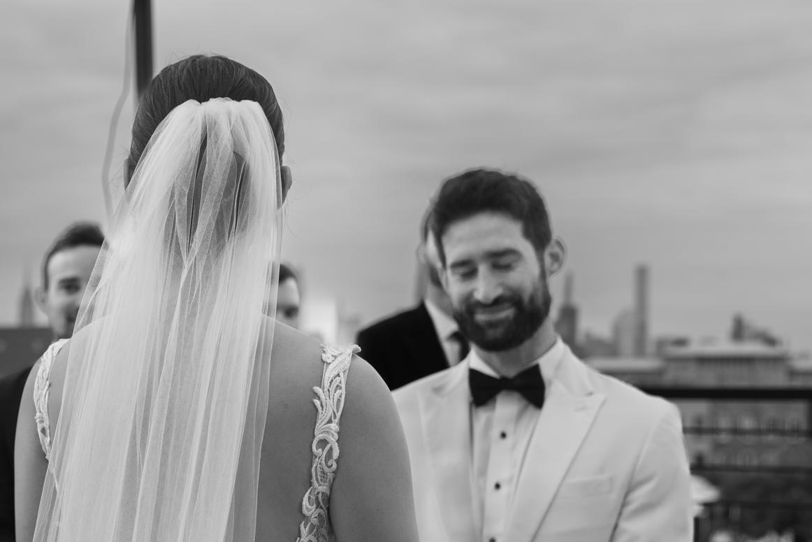 Groom smiling during the vows at a rooftop ceremony with the city skyline behind him