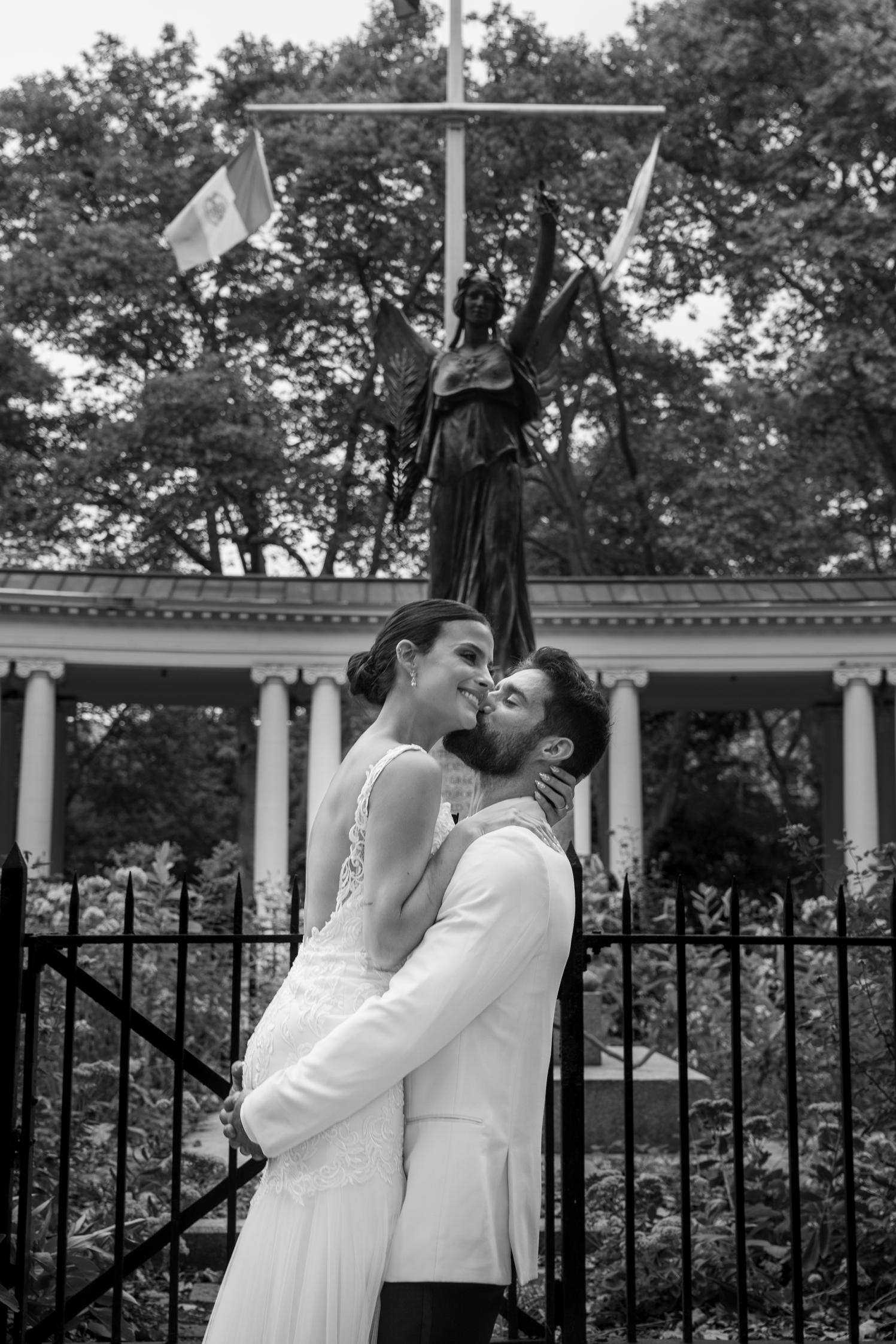 Groom kissing his bride's cheek beneath a winged bronze statue