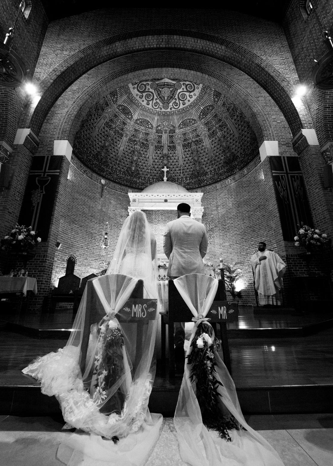 Bride and groom kneeling at the altar of a brick church, MR and MRS signs on their chairs